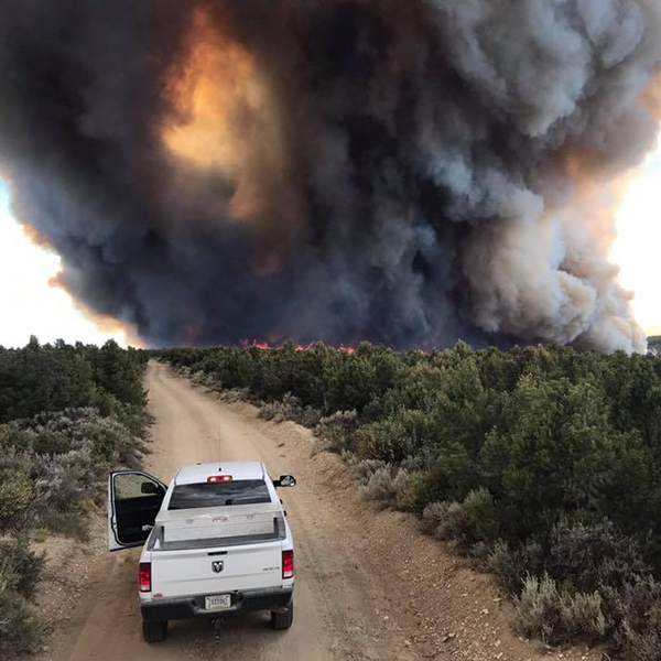 Smoke billows in the background as a fire burns. White truck from fire department in foreground.